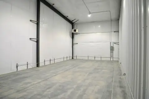 Inside an empty SYT Storage unit with a concrete floor, white-lined walls, lighting, outlets and a ceiling-mounted gas heater